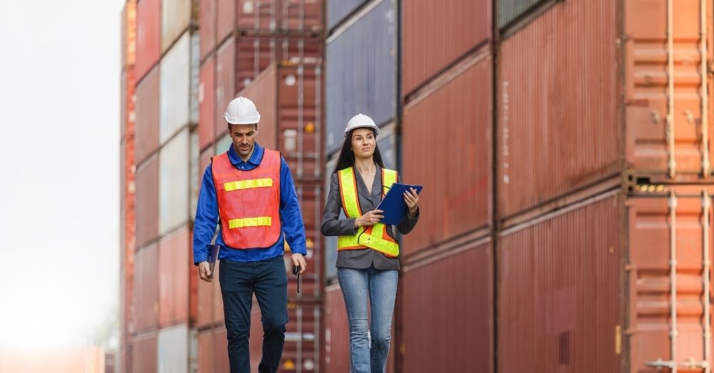 Two port workers walk past stacked shipping containers.