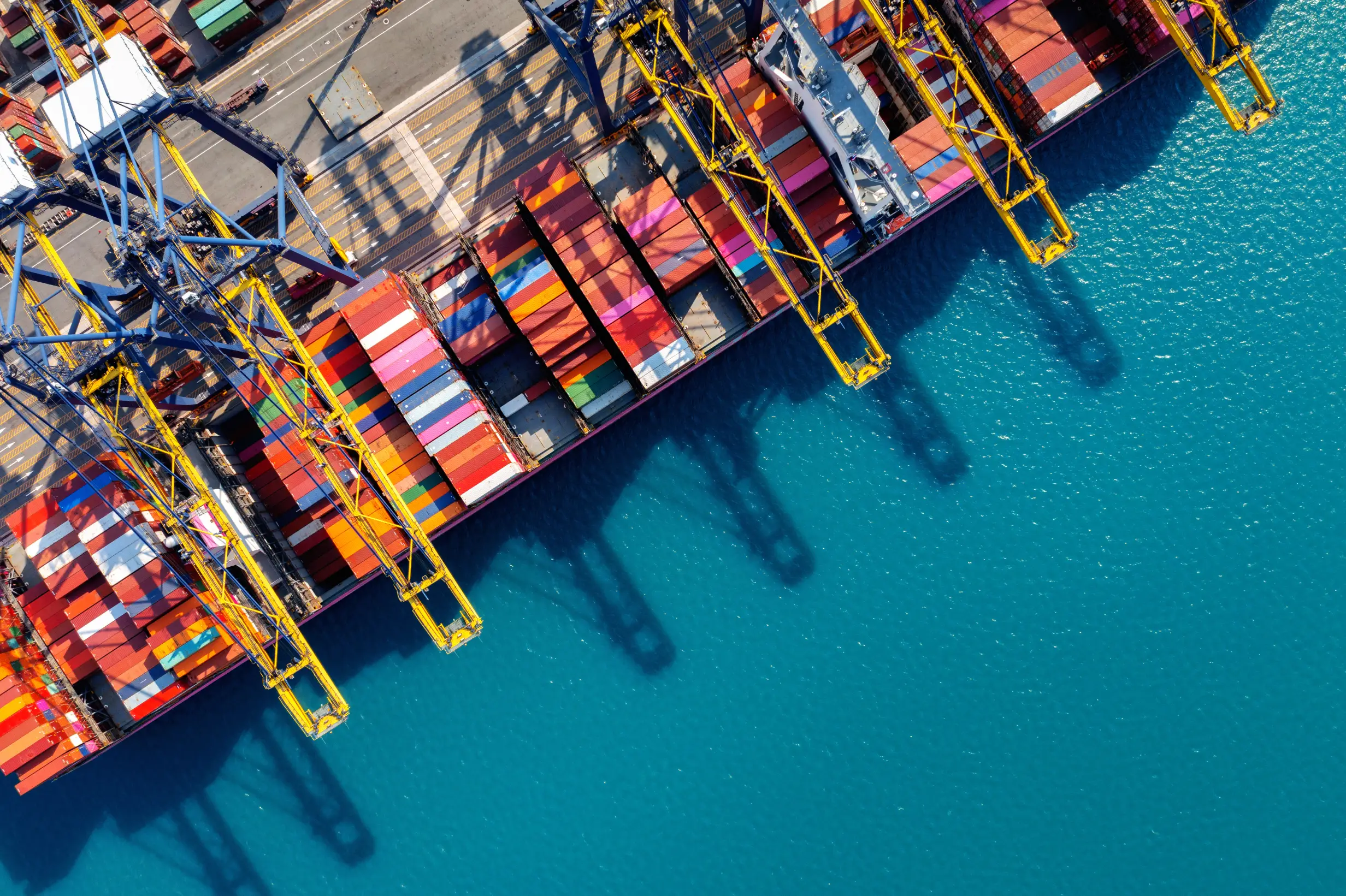 Top-down aerial view of a cargo ship docked at a port, loaded with colorful shipping containers and surrounded by large yellow and blue cranes above turquoise harbor water