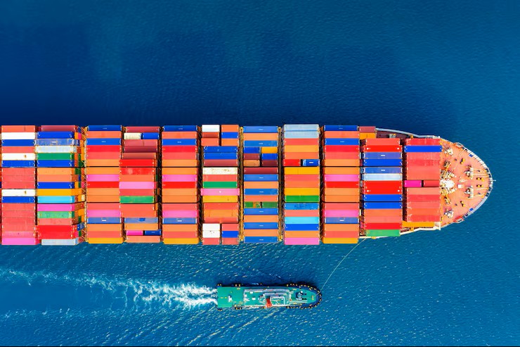 Aerial view of a large container ship loaded with multicolored shipping containers escorted by a tugboat on blue water — drayage in logistics.